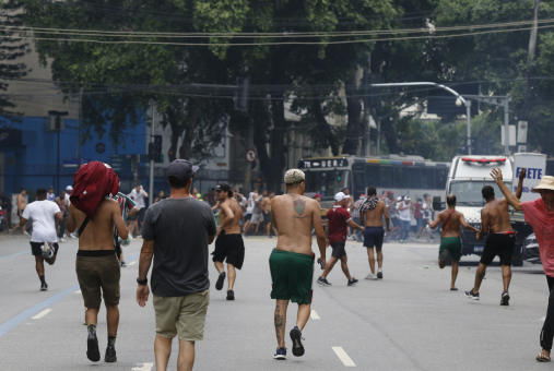 Viewed from behind, Fluminense fans wearing team shirts and some shirtless individuals are seen rushing towards a conflict. In the background, a group is engaged in a collective brawl outside Maracanã stadium in Rio de Janeiro