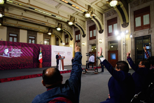 Periodistas en primer plano levantando la mano para preguntar en una conferencia de prensa. Al fondo se ve el presidente de México en un atril