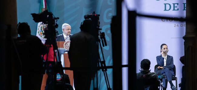 President Andrés Manuel López Obrador, of Mexico, talks surrounded by shadows and cameras; to the right, Claudia Sheinbaum is seated