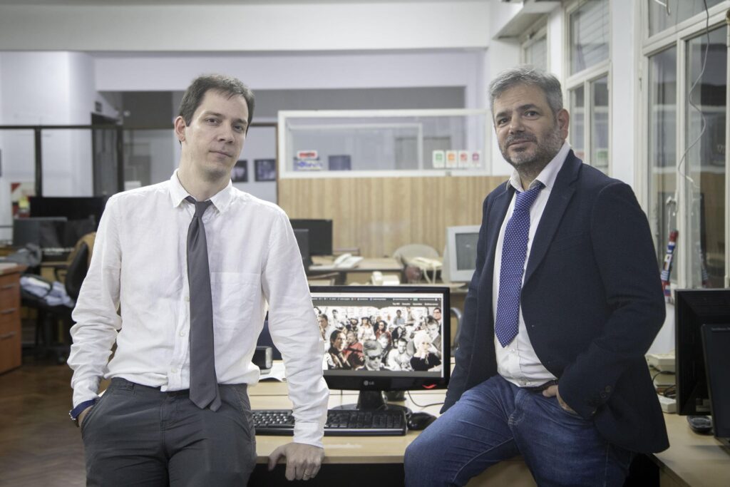 A frontal photo of Federico Poore and Manuel Barrientos, the Argentinian journalists behind "Periodistas en el Cine," wearing suits. Between them, there's a computer with a screen showing the "Periodistas en el Cine" banner.