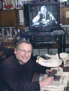 A portrait of Joe Saltzman, the founder of the Image of the Journalist in Popular Culture database. In the background, there's a TV showing a black and white movie with a journalist speaking on the phone
