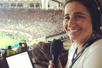 A woman wearing headphones in front of a computer screen smiling inside a soccer stadium