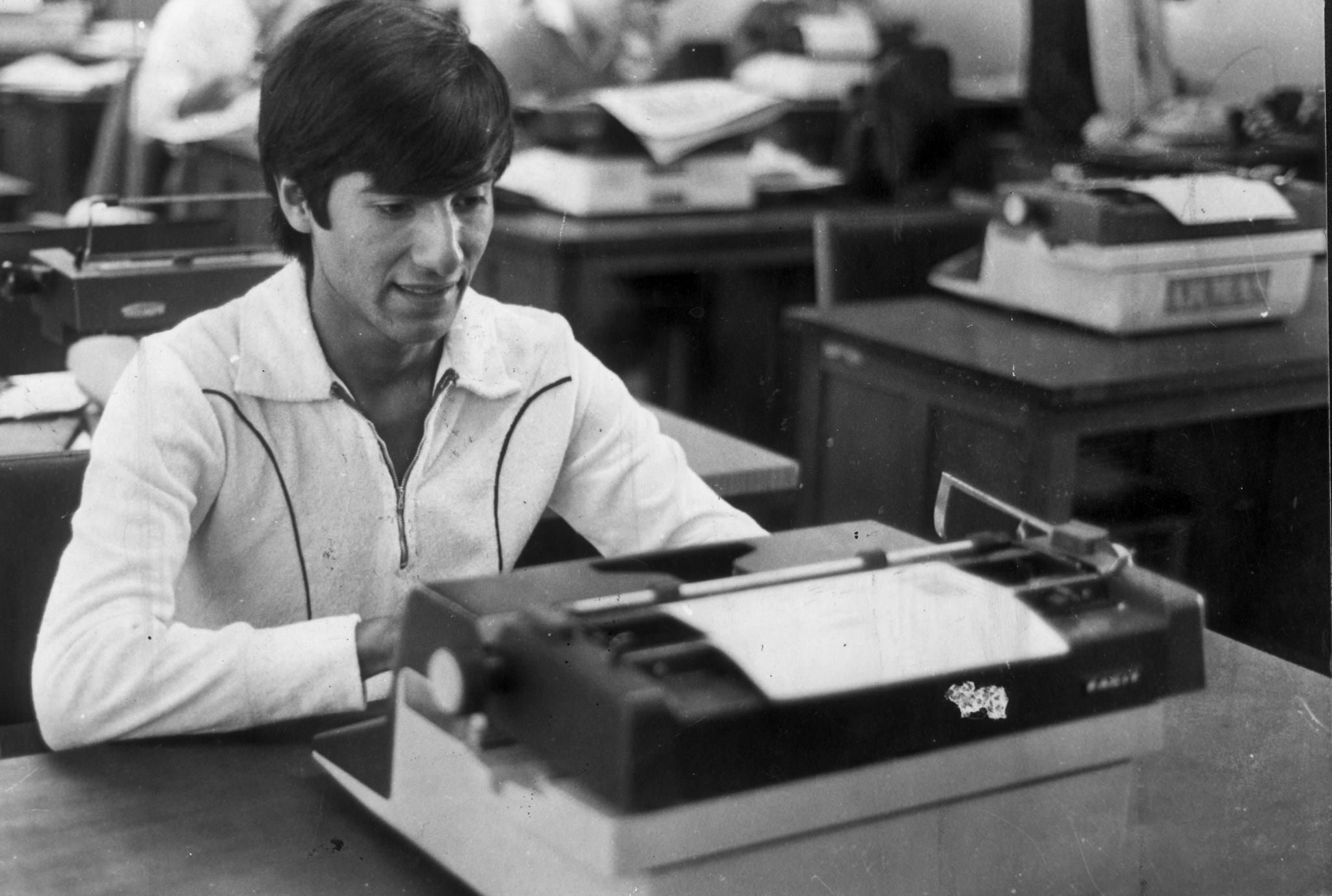 Foto en blanco y negro de un joven periodista sentado frente a su escritorio escribiendo en su máquina de escribir mientras sonríe. 