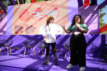 Two women are standing on a stage during a journalism festival. One, on the left, is wearing sunglasses and a white shirt, while the other, on the right, is holding a microphone and wearing a colorful shirt and black pants.