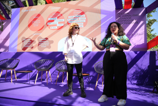 Two women are standing on a stage during a journalism festival. One, on the left, is wearing sunglasses and a white shirt, while the other, on the right, is holding a microphone and wearing a colorful shirt and black pants.