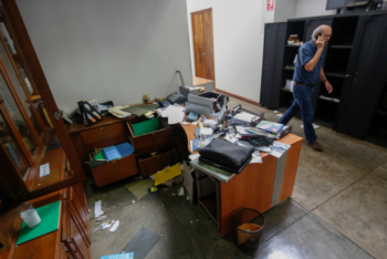 Photo of a disorganized office with papers, files, and office supplies scattered across desks and the floor. A man is seen in the background talking on the phone, walking past cabinets and desks in disarray.