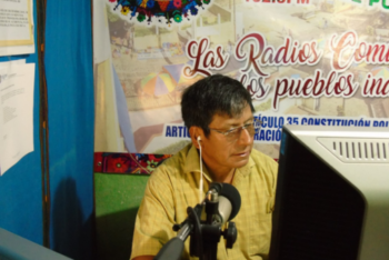 A man, Anselmo Xunic from Cultural Survival, is inside a radio studio, sitting in front of a microphone. Behind him, a banner reads "las radios comunitarias, los pueblos indígenas."