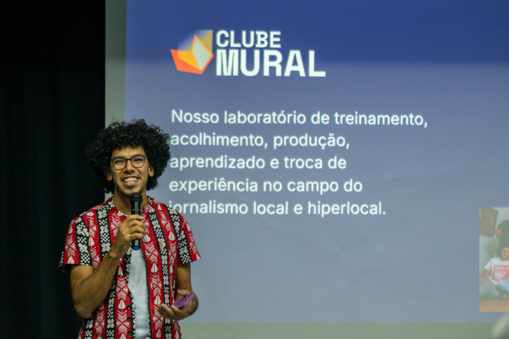 A man with curly hair, glasses, and a bright smile speaks into a microphone on stage. He wears a red patterned shirt over a white t-shirt