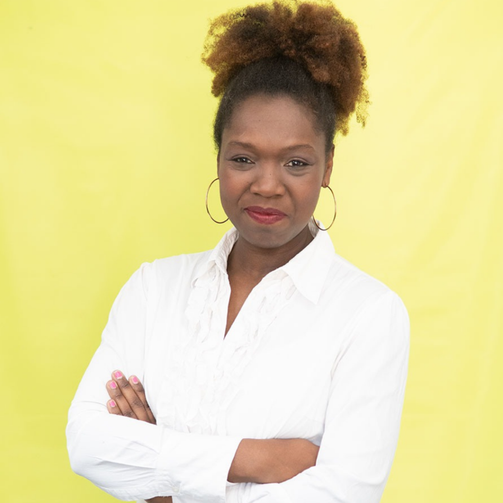 A woman with a confident expression stands against a yellow background, arms crossed.