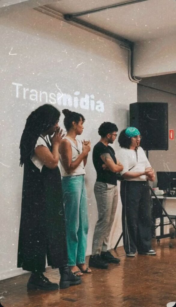 Four individuals stand in a row on a wooden floor, each engaged in discussion or contemplation, in front of a projection that reads 'Transmídia.'