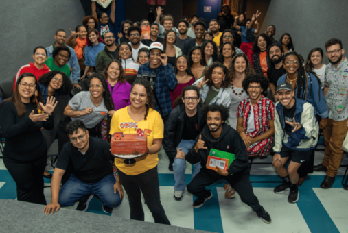 A large group of people smiling and posing for a group photo in a brightly lit room. At the front, a woman in a yellow shirt holds a decorative cake, surrounded by others making cheerful gestures.