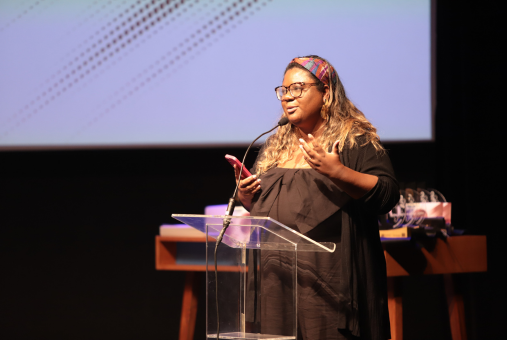 A woman with glasses, wearing a black outfit and a colorful headband, speaks into a microphone at a podium on stage