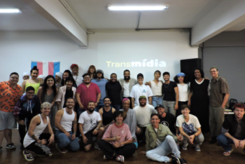 A diverse group of people poses for a group photo in a room with bright lighting. Behind them, a screen displays the word 'Transmídia,' and a transgender pride flag is visible on the wall.