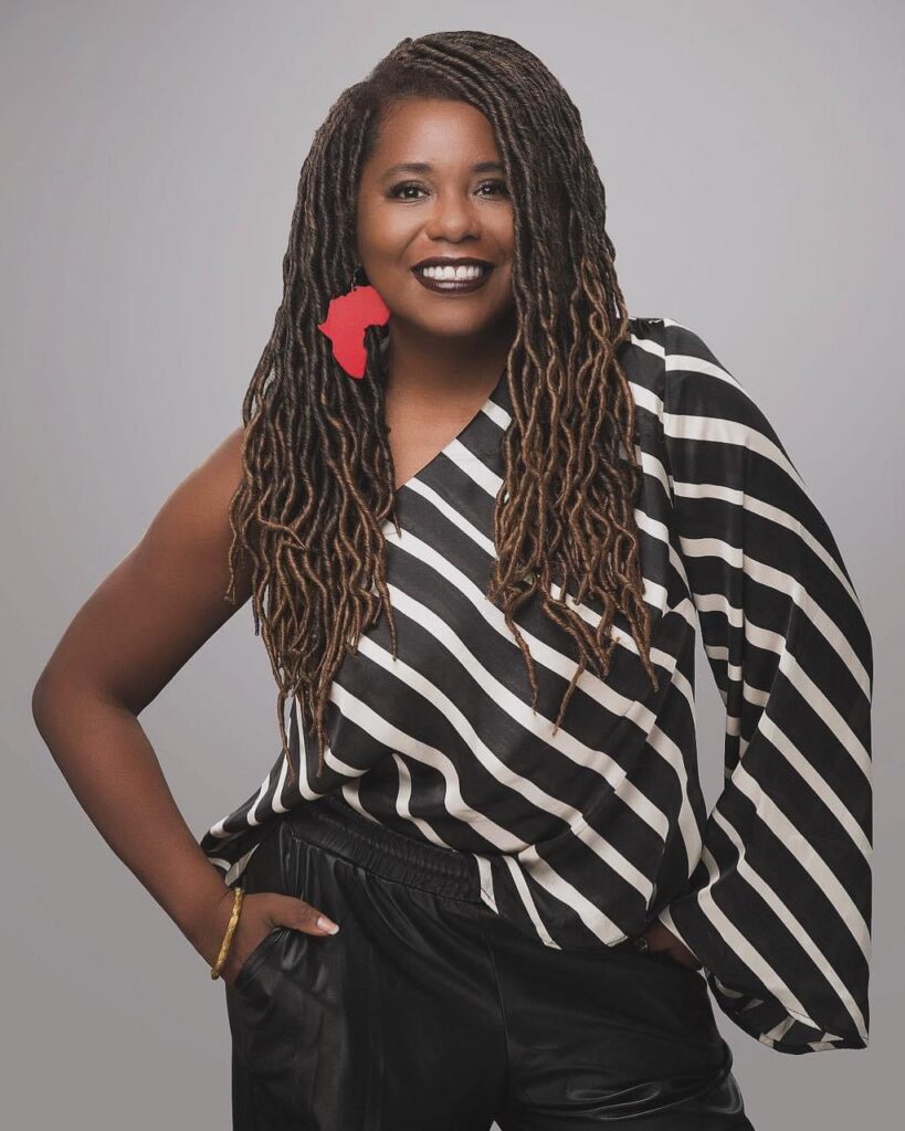 A woman with long locs and a warm smile stands with one hand in her pocket against a neutral background. She wears a black and white striped, one-shoulder blouse, black pants, and a bold red earring shaped like the African continent.