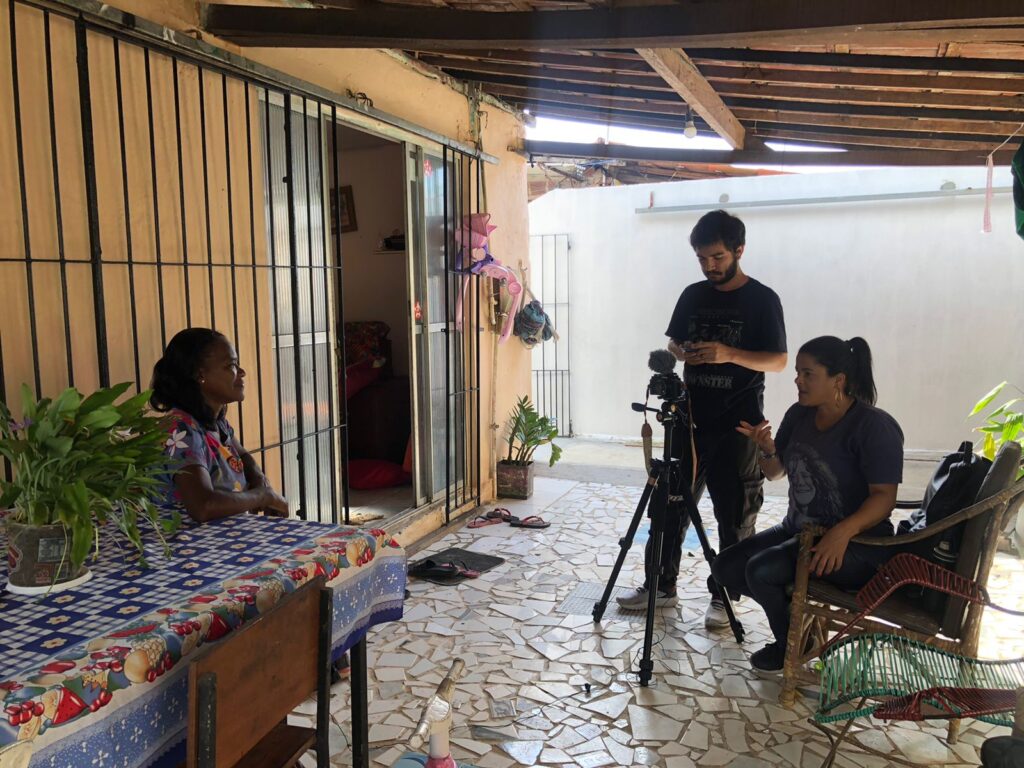 Uma mulher sentada à mesa decorada com uma toalha azul e vermelha, sendo entrevistada por uma jornalista e um cinegrafista. A cena ocorre em uma varanda com piso de azulejo, telhado de madeira, plantas em vasos e atmosfera descontraída. A jornalista gesticula enquanto o cinegrafista ajusta o equipamento.