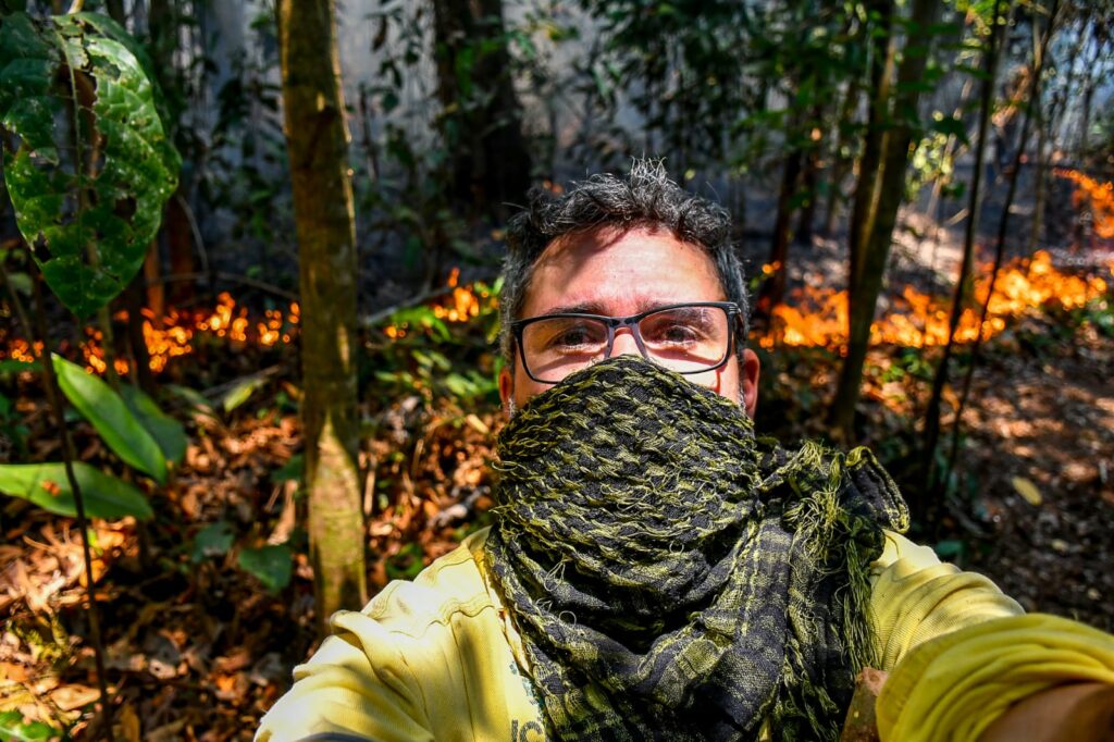 Uma selfie de um jornalista usando óculos e uma echarpe cobrindo o rosto, em pé em uma floresta. O fundo mostra chamas ativas e vegetação densa. Ele está documentando os esforços de combate às queimadas em território indígena.