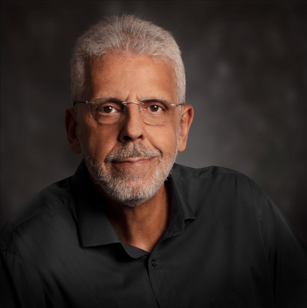 Portrait of Ernesto Rodrigues, author, journalist, and journalism professor, with short white hair, glasses, and a trimmed beard, wearing a dark shirt