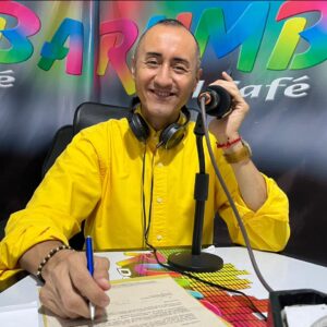 Hombre de mediana edad sentado en una mesa con un micrófono en mano y audífonos en su cuello sonriendo a la cámara