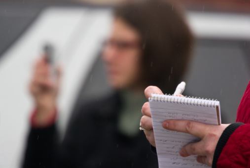 A close-up of a journalist’s hand holding a notebook and pen, with blurred background showing another person holding a smartphone