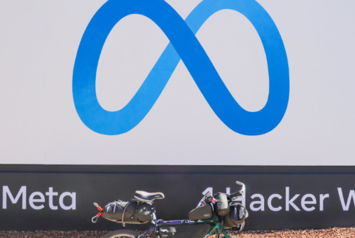 bicycle parked in front of a large Meta sign at the company's headquarters in Menlo Park. The sign displays the Meta logo (a blue infinity symbol) and the phrase "Meta - 1 Hacker Way," surrounded by trees and greenery.