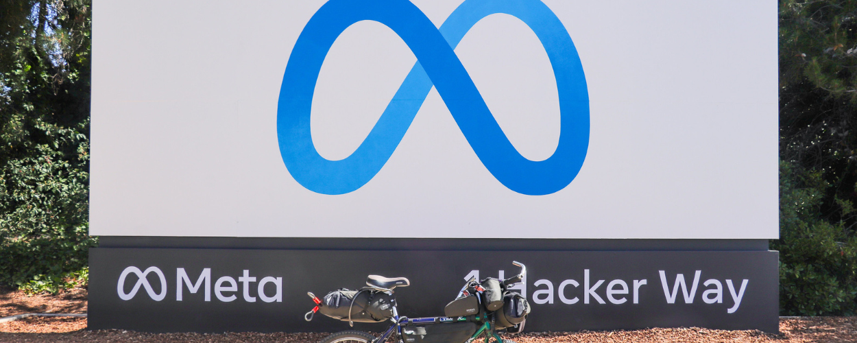 bicycle parked in front of a large Meta sign at the company's headquarters in Menlo Park. The sign displays the Meta logo (a blue infinity symbol) and the phrase "Meta - 1 Hacker Way," surrounded by trees and greenery.