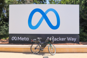 bicycle parked in front of a large Meta sign at the company's headquarters in Menlo Park. The sign displays the Meta logo (a blue infinity symbol) and the phrase "Meta - 1 Hacker Way," surrounded by trees and greenery.