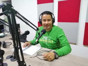 Hombre joven en una cabina de radio, sentado frente a una mesa usando audífonos y un micrófono sonriendo a la cámara