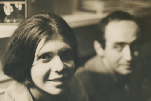 Black-and-white photograph of Clarice and Vladimir Herzog. Clarice, in the foreground, smiles gently at the camera, while Vladimir, slightly out of focus in the background, looks in her direction. The setting appears to be an indoor space with bookshelves behind them.