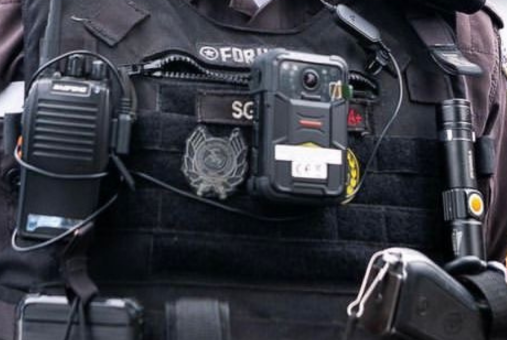 A close-up of a police officer's tactical vest featuring a body camera securely attached to the chest. The vest also holds a radio communication device, a flashlight, and other tactical gear. The officer's uniform includes identification badges and insignia.