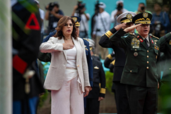 Rixi Moncada, presidential candidate and current Minister of Defense of Honduras, stands with her right hand over her heart, wearing a beige suit. Beside her, General Hernández, in a green military uniform with numerous medals, salutes. In the background, other military personnel and photographers are visible.