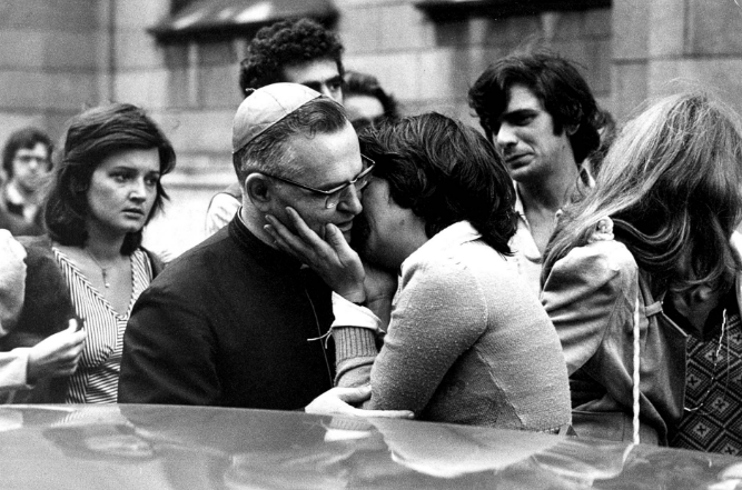 Black-and-white photograph of Clarice Herzog and Cardinal Dom Paulo Evaristo Arns leaving the ecumenical service at São Paulo’s Sé Cathedral on October 31, 1975. Clarice, visibly emotional, walks alongside the archbishop, who played a key role in denouncing the crimes of Brazil’s military dictatorship. The service was held in memory of her husband, journalist Vladimir Herzog, murdered days earlier.