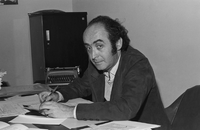 Black-and-white photograph of journalist Vladimir Herzog sitting at his desk at TV Cultura on October 9, 1975. He holds a pen and looks at the camera with a serious expression. Papers are spread across the desk, and a typewriter is visible in the background. This image was taken just weeks before his assassination by Brazil’s military dictatorship.