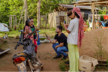 Journalists interview an elderly woman in a rural community in the Amazon. The scene takes place outdoors, with wooden houses and hanging clothes in the background. One journalist is kneeling, holding a recorder, while another films with a cellphone.