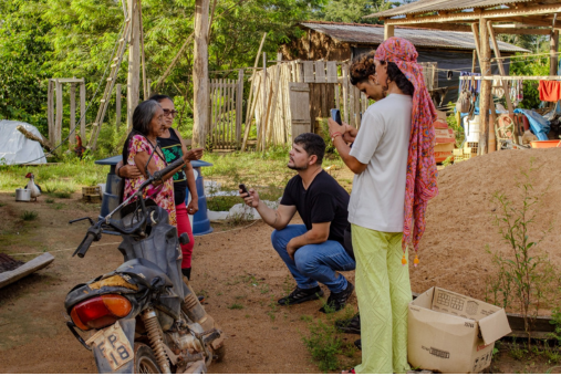 Journalists interview an elderly woman in a rural community in the Amazon. The scene takes place outdoors, with wooden houses and hanging clothes in the background. One journalist is kneeling, holding a recorder, while another films with a cellphone.