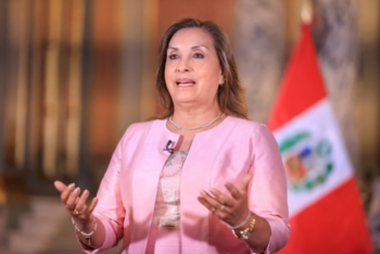 Dina Boluarte speaking during a public address, wearing a pink jacket, with a Peruvian flag behind her.