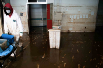 A person wearing protective gear and a face mask walks through a flooded and mud-covered corridor inside a building, likely affected by recent heavy rains. Water marks are visible on the walls, and debris is scattered across the floor.