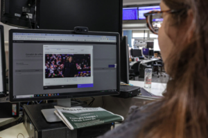 A woman is sitting at a desk in a newsroom, looking at a computer monitor. The screen displays a video editing tool with a paused video of a woman speaking on stage. 