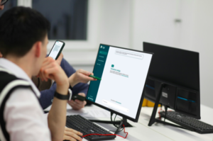 Três pessoas sentadas em frente a computadores observam e discutem a interface de um chatbot exibida em uma das telas. 
