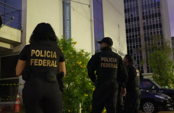 : Three Federal Police officers, seen from behind, stand in front of a Brazilian Social Security (INSS) office.