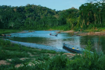 Vista de um rio cercado por floresta densa na Amazônia, com dois barcos pequenos navegando e uma canoa estacionada na margem.