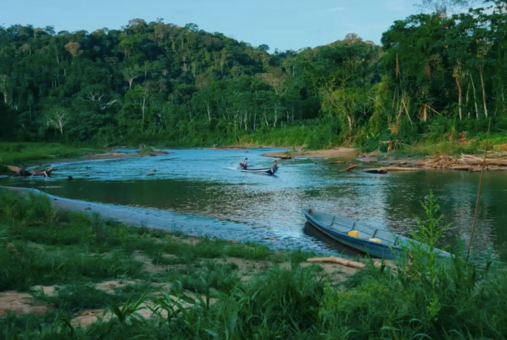 Vista de um rio cercado por floresta densa na Amazônia, com dois barcos pequenos navegando e uma canoa estacionada na margem.