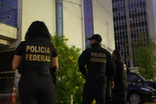 Three Federal Police officers, seen from behind, stand in front of a Brazilian Social Security (INSS) office.