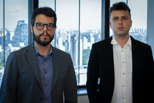 Two journalists pose in front of a window overlooking the city. On the left is Fábio Leite, editor of Metrópoles in São Paulo. On the right is Luiz Vassallo, investigative reporter at Metrópoles