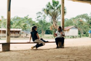 Duas pessoas conversam sentadas em uma área aberta de uma comunidade amazônica, com casas de madeira ao fundo e vegetação típica da floresta.