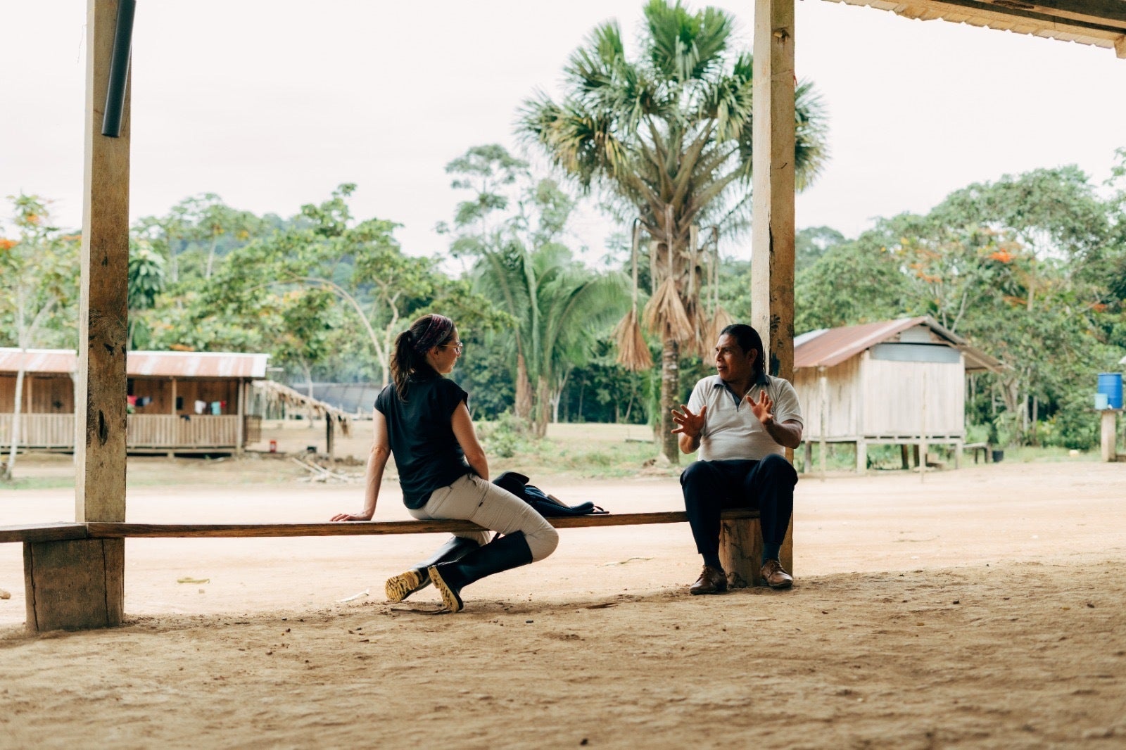 Duas pessoas conversam sentadas em uma área aberta de uma comunidade amazônica, com casas de madeira ao fundo e vegetação típica da floresta.