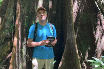 Dom Phillips em pé na floresta amazônica, com boné, camiseta azul e mochila, segurando um caderno, posicionado entre as raízes de uma grande árvore.