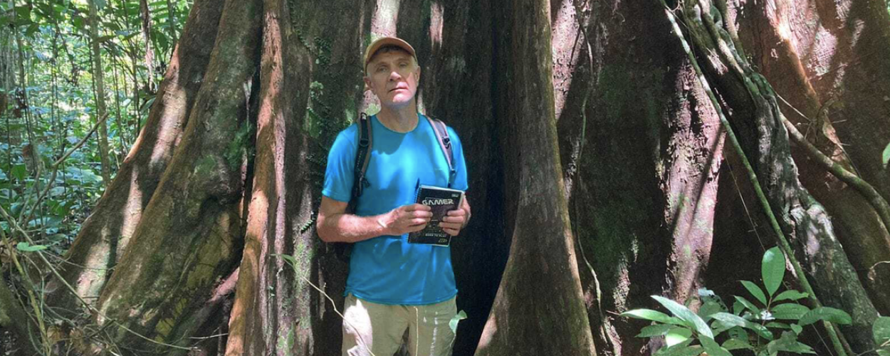 Dom Phillips em pé na floresta amazônica, com boné, camiseta azul e mochila, segurando um caderno, posicionado entre as raízes de uma grande árvore.