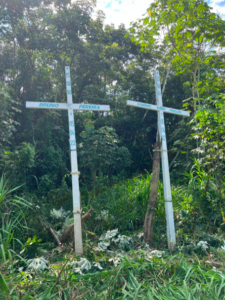 Duas cruzes brancas fincadas na mata, com os nomes de Bruno Pereira e Dom Phillips, além das datas de nascimento e morte. Ao fundo, vegetação densa da floresta amazônica.