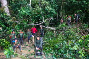 Grupo de cerca de 12 pessoas, majoritariamente homens indígenas, posando na floresta amazônica, cercados por vegetação densa e galhos caídos. Alguns estão em pé, outros sentados sobre troncos, todos com roupas apropriadas para expedição na selva, mochilas e botas. Ao fundo, mata fechada.
