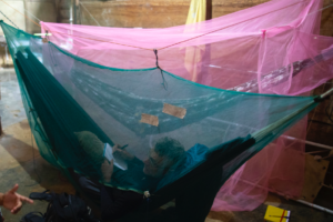 Pessoa deitada em uma rede protegida por um mosquiteiro verde, escrevendo em um caderno com uma lanterna de cabeça acesa. Atrás dela, há outra rede coberta por um mosquiteiro rosa. No chão, ao lado da rede, estão uma mochila, chinelos e um caderno amarelo. O ambiente tem piso de cimento e paredes de madeira, típico de alojamentos simples na região amazônica.